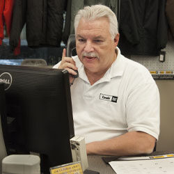 A man wearing a white Carolina Cat polo shirt sits at a desk looking at a computer while on the phone.